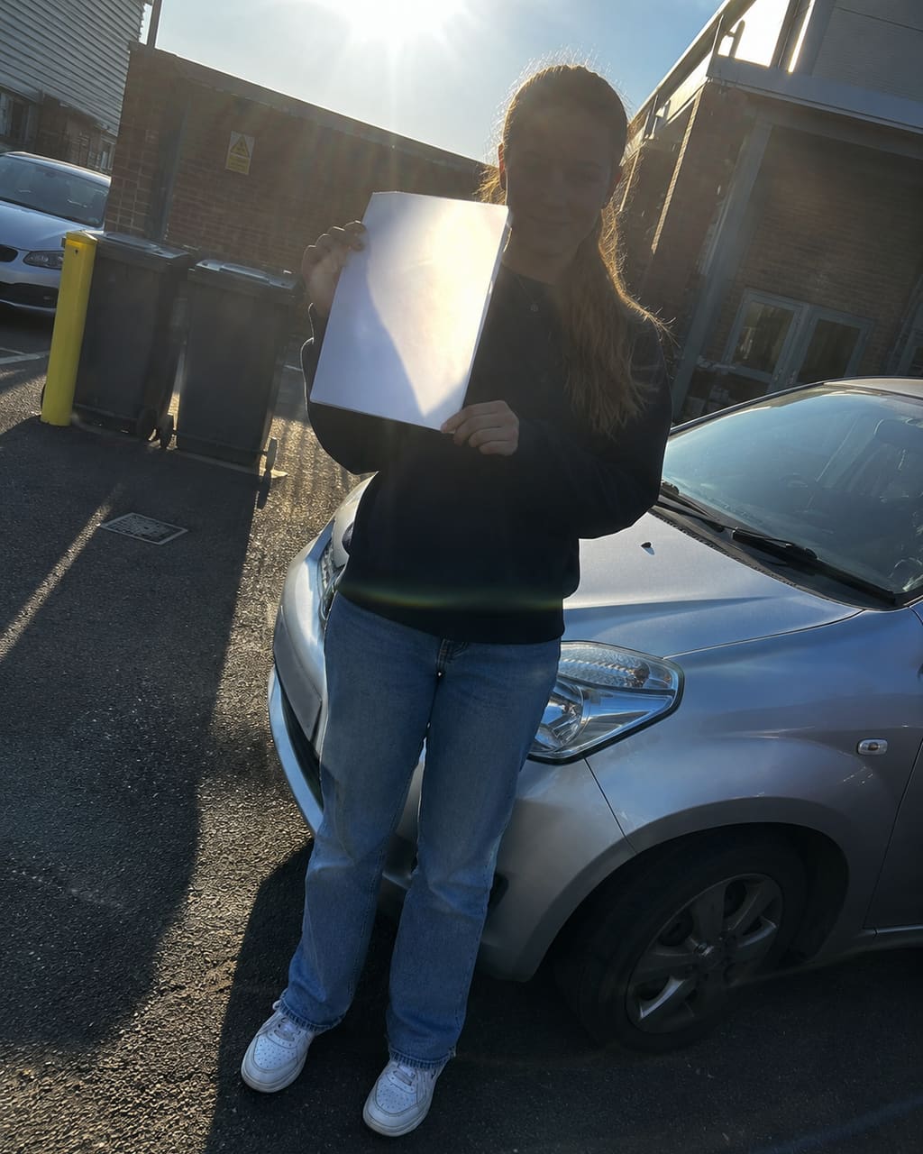 The photograph most instructors take: a pupil holding a pass certificate beside the car in a test centre car park, shot into the sun, roof line cut off, bins behind them.