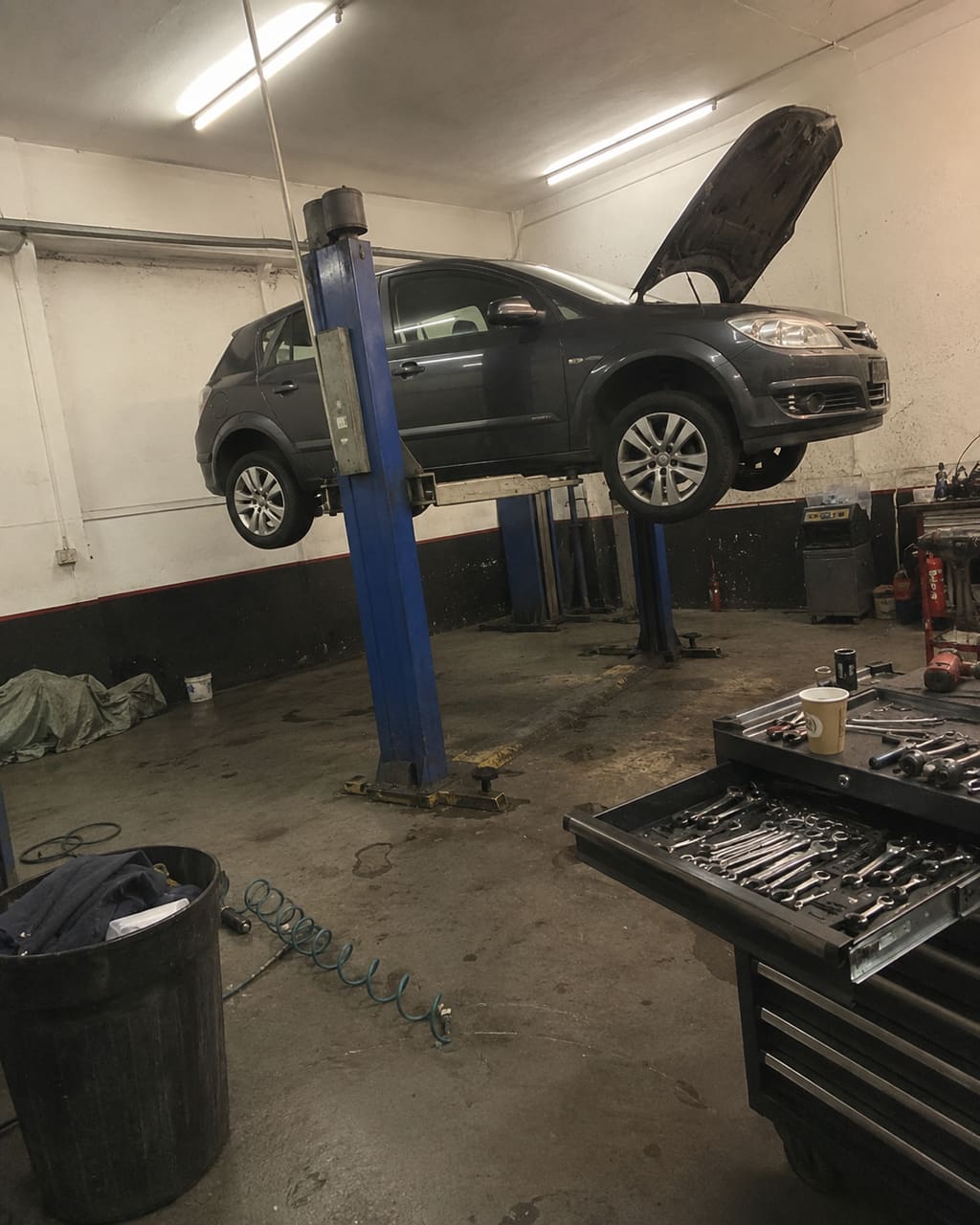 The photograph most garages take: a car on the ramp shot from the workshop floor under strip lights, a bin, an air line and half a toolbox in the frame.