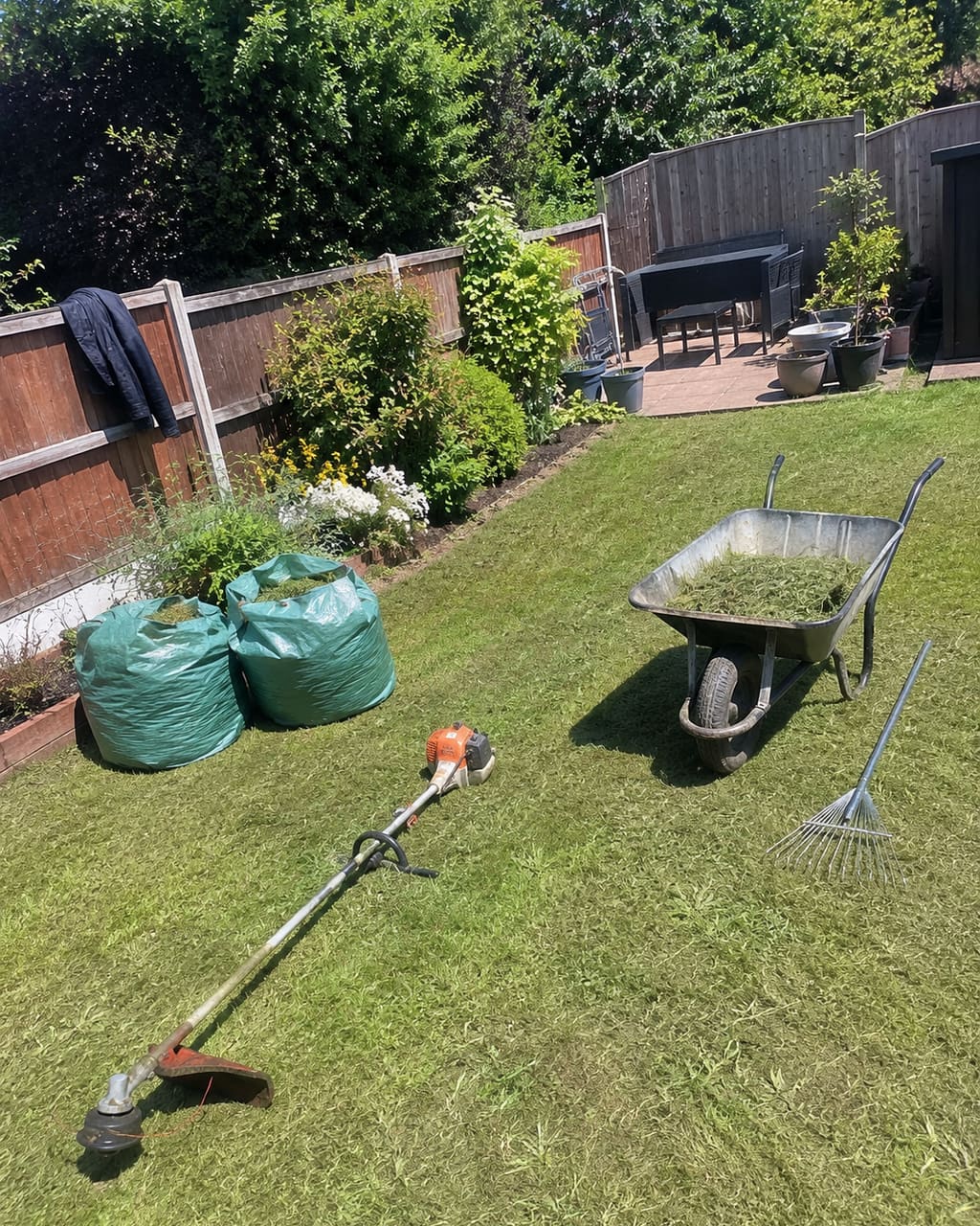 The photograph most gardeners take: a finished lawn and border shot from the middle of the grass at midday, sun overhead, half a wheelbarrow in the corner.