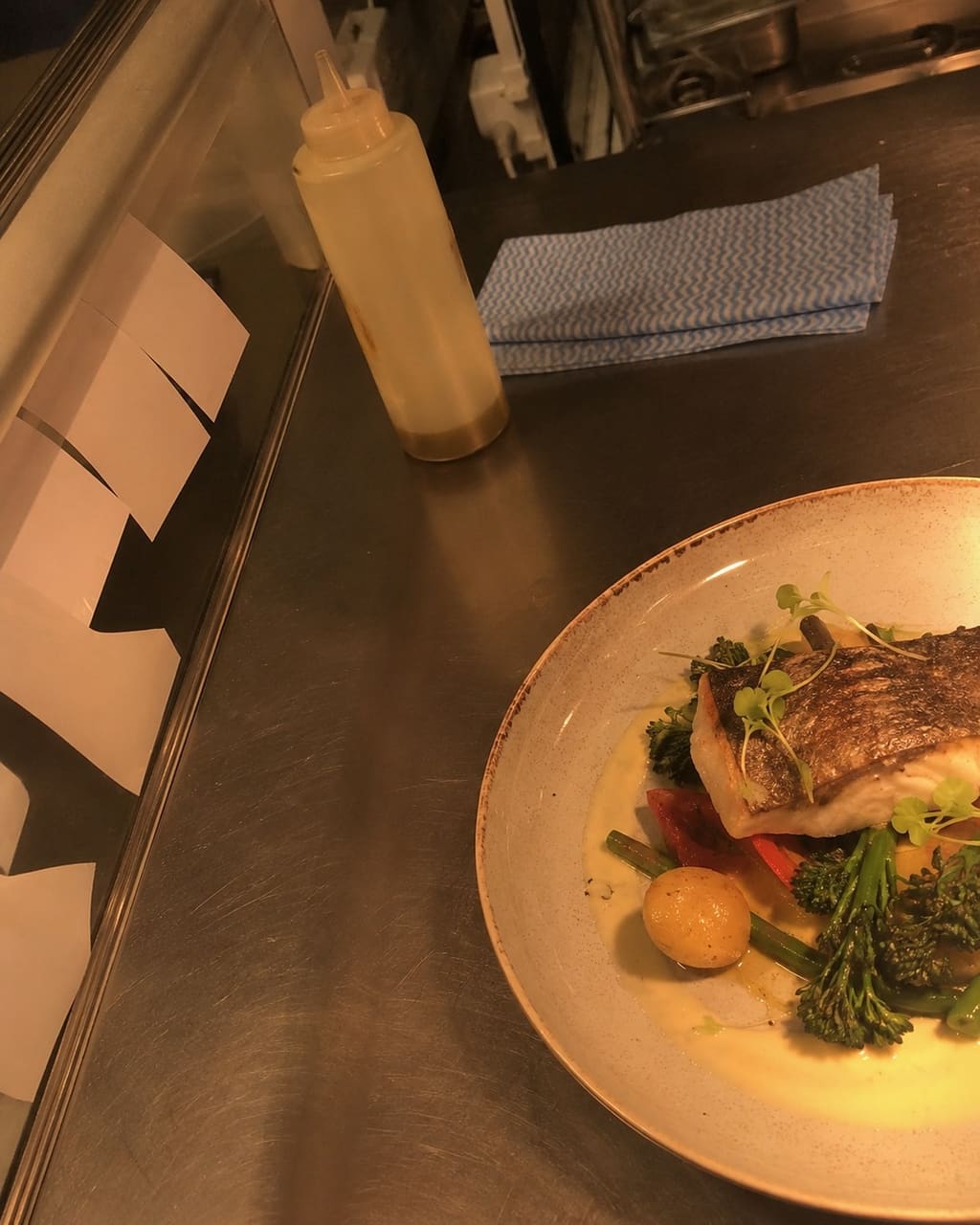 The photograph most kitchens take: a plated dish shot from above during service under orange overhead lighting, a check and a cloth in the corner of the frame.